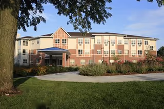Exterior view of a multi-story assisted living facility building with beige and brick facade, surrounded by green grass, bushes, and a large tree under a clear blue sky.