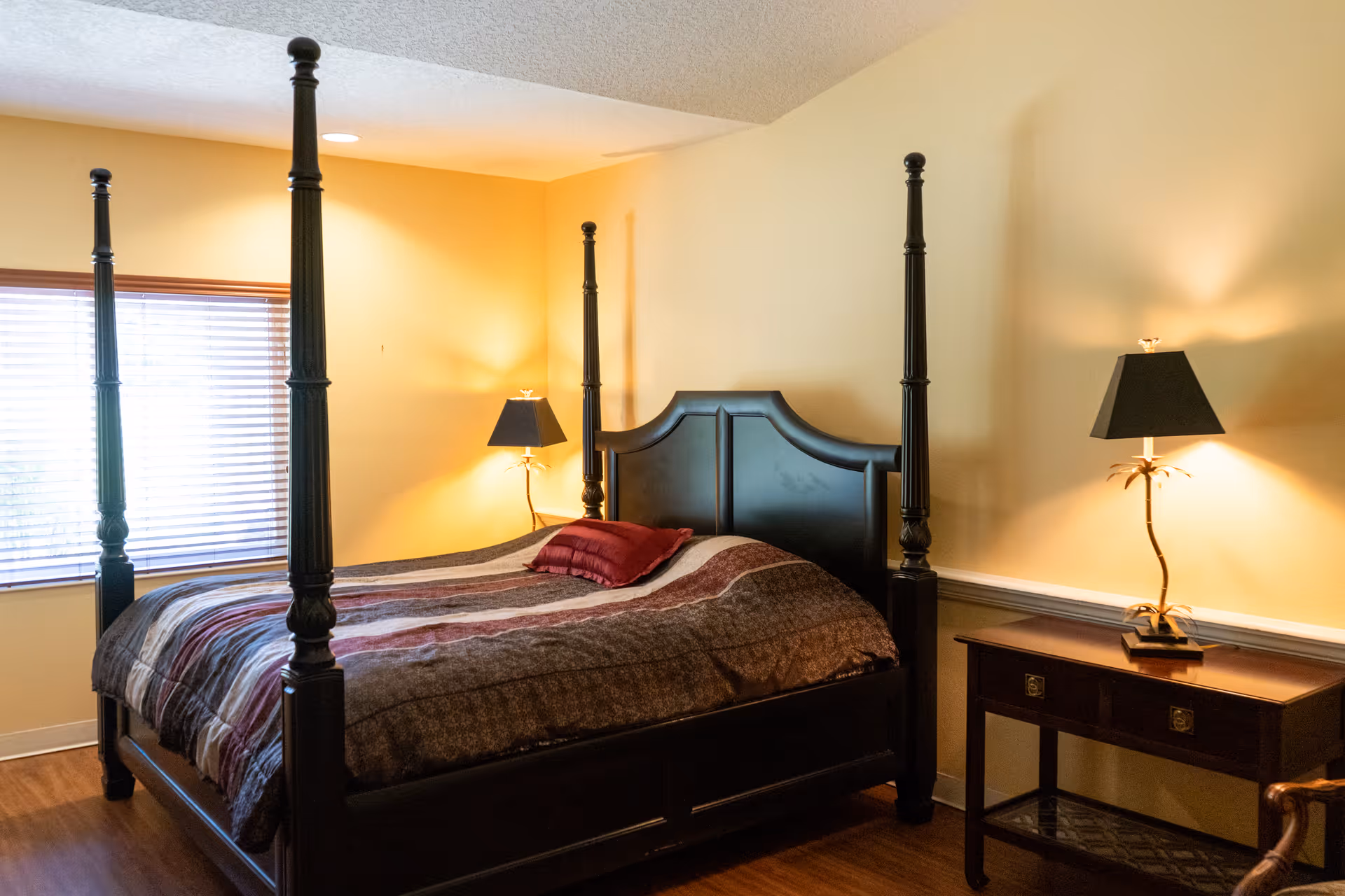 A bedroom with a dark wooden four-poster bed featuring a striped bedspread and a red pillow. There are two matching bedside tables, each with a lamp that has a black shade and a decorative base. The room has light yellow walls and a window with wooden blinds letting in natural light.