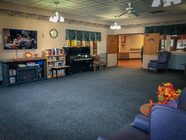 A spacious common area in a senior living facility with carpeted floor, a piano, bookshelves filled with books, a wall-mounted TV showing a scene from a movie, and comfortable chairs. The room has a ceiling fan, hanging light fixtures, and an open doorway leading to another room with a cross on the wall.