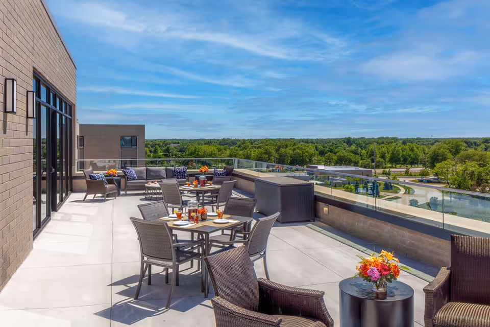 A spacious outdoor patio area with multiple tables and chairs arranged for seating. The patio features glass railings providing a clear view of green trees and a blue sky. There are flower arrangements on the tables and comfortable cushioned seating along the far side near the building wall.