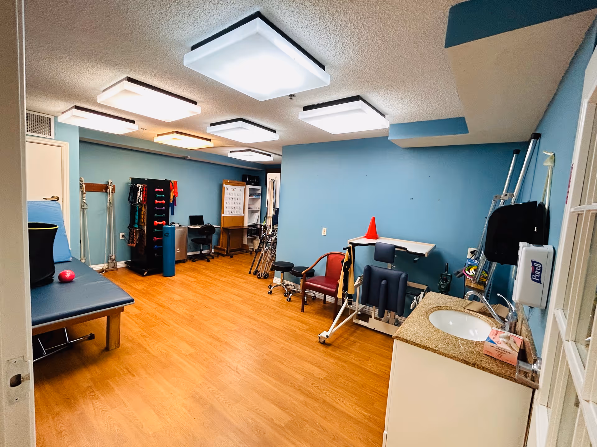A therapy or rehabilitation room with light blue walls and wooden flooring. The room contains various physical therapy equipment including a padded therapy table, resistance bands, weights, a small desk with a chair, a red chair, and other assistive devices. There is a sink with a Purell hand sanitizer dispenser mounted on the wall nearby. The ceiling has multiple square light fixtures.