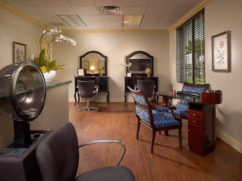 Interior view of a salon area in a senior living facility featuring two styling stations with mirrors and chairs, a hair dryer chair in the foreground, a manicure table with two blue upholstered chairs, wooden flooring, and a window with blinds letting in natural light.