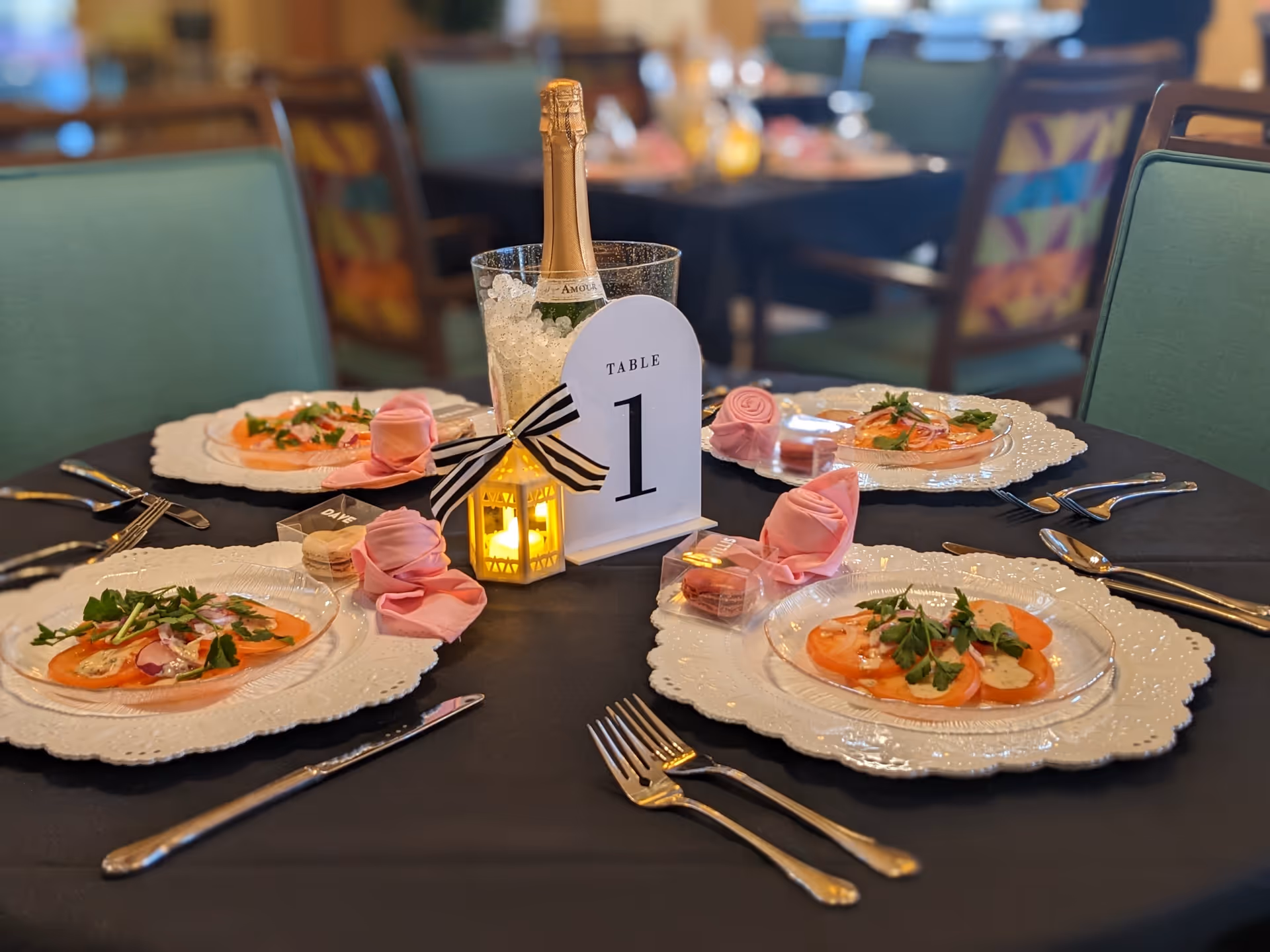 A dining table set for four with white decorative plates holding a salad of sliced tomatoes and greens. Each place setting has a pink cloth napkin folded into a rose shape, silver cutlery, and a small clear box with macarons. In the center of the table is a bottle of champagne in an ice bucket, a small lit lantern with a black and white striped bow, and a table number sign reading 'Table 1'. The chairs around the table have green upholstery, and the background shows more tables and chairs in a softly lit dining area.