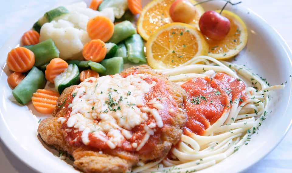 A plate of food featuring spaghetti with tomato sauce, a breaded chicken cutlet topped with melted cheese and tomato sauce, steamed mixed vegetables including carrots, green beans, cauliflower, and zucchini, garnished with two orange slices and a cherry on top.