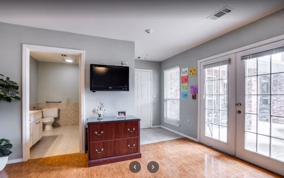 Bright common living area with a wall-mounted TV above a wooden dresser, an open bathroom doorway, and glass French doors to the outside.