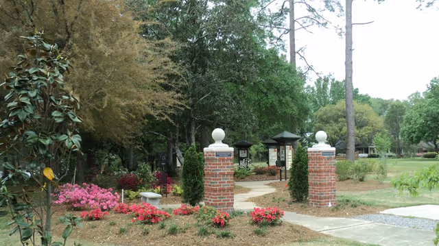 A landscaped outdoor area at St. Joseph Assisted Living Center featuring two brick pillars with white spherical tops at the entrance of a winding concrete pathway. The pathway is surrounded by green trees, bushes, and pink flowering plants, with a bench and informational signs visible along the path.