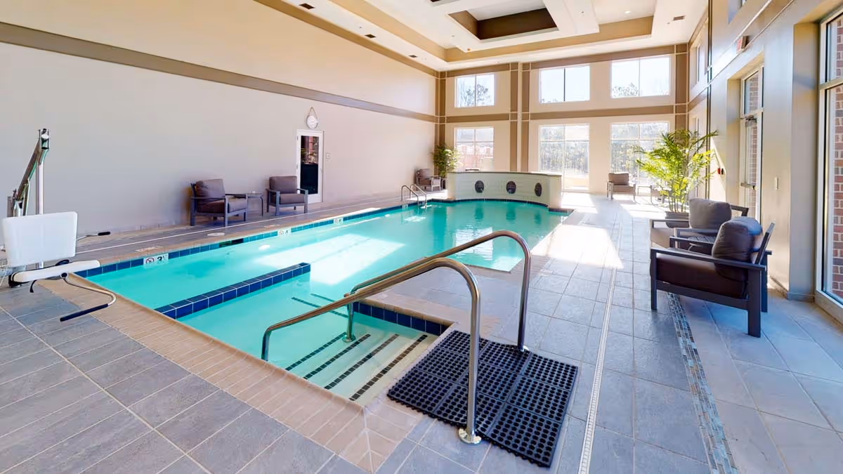 Indoor swimming pool area with clear blue water, surrounded by tiled flooring and several cushioned chairs along the walls. Large windows allow natural light to fill the space, and there are plants near the windows. A pool lift is installed for accessibility.