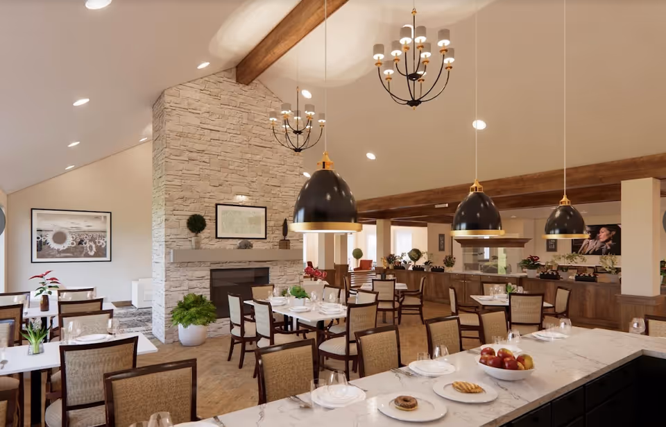 A spacious dining room in Cedarhurst Senior Living of Topeka featuring multiple tables with chairs, set with plates, glasses, and cutlery. The room has a high ceiling with wooden beams, three large black pendant lights, and two chandeliers. A stone fireplace with framed artwork above it is visible, along with various plants and decorative items. The overall ambiance is warm and inviting.