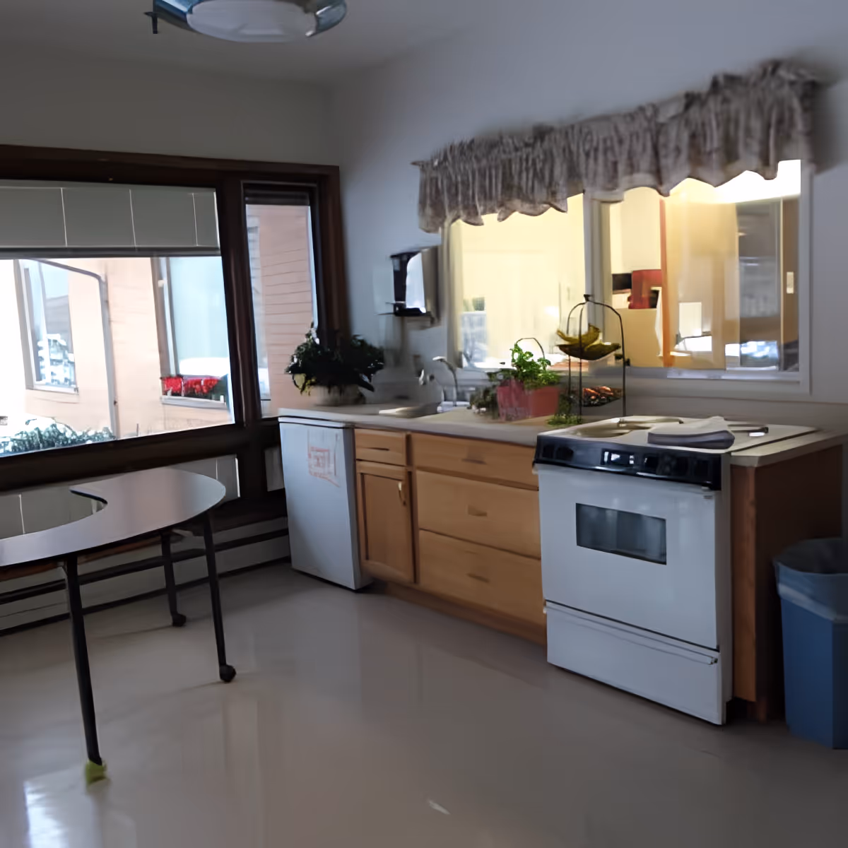 Small kitchen area with a stove, sink, wooden cabinets and a round table by large windows.