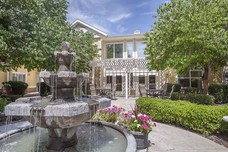 Outdoor courtyard area at Brookdale Boise Parkcenter featuring a multi-tiered stone water fountain surrounded by a circular pool. There are green bushes, colorful flowers in pots, and several trees providing shade. In the background, there is a yellow building with large windows and a white pergola over patio tables and chairs.