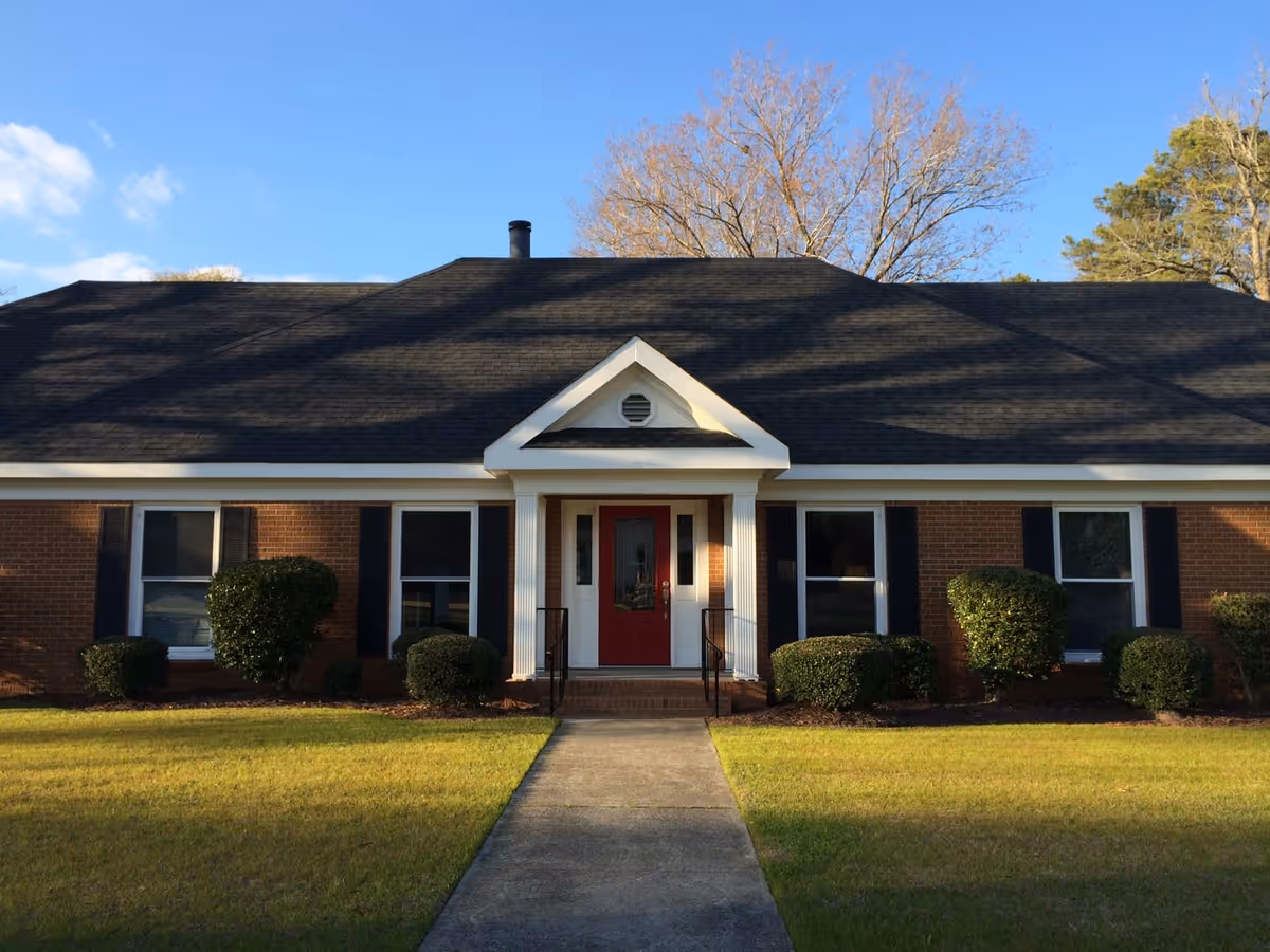 Front exterior view of a single-story brick building with a black shingled roof, a red front door with white trim, and four windows with black shutters. There are neatly trimmed bushes in front of the building and a concrete walkway leading to the entrance. Trees are visible in the background under a blue sky with some clouds.