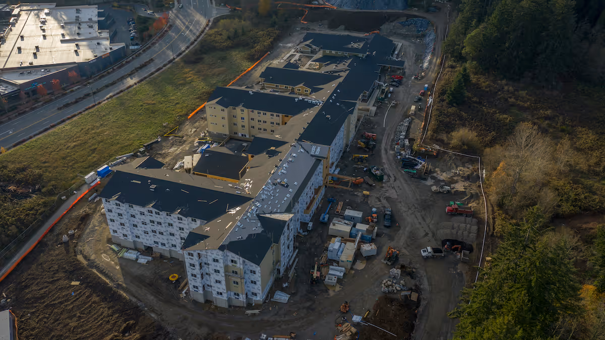 Aerial view of a large building under construction surrounded by dirt roads, construction equipment, and materials. The building has multiple wings and is partially covered with white and yellow exterior sheathing. The surrounding area includes a grassy field, a road, and a wooded area.