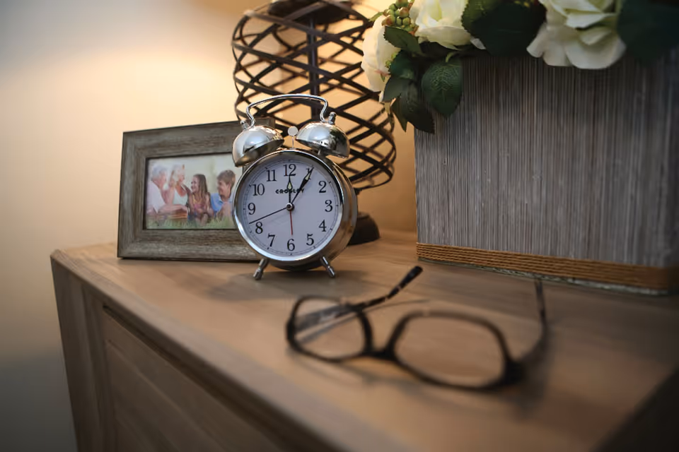 A wooden surface with a silver analog alarm clock showing 12:05, a framed photo of four people smiling, a decorative lamp with a black wireframe base, a large gray planter with white flowers, and a pair of black eyeglasses in the foreground.