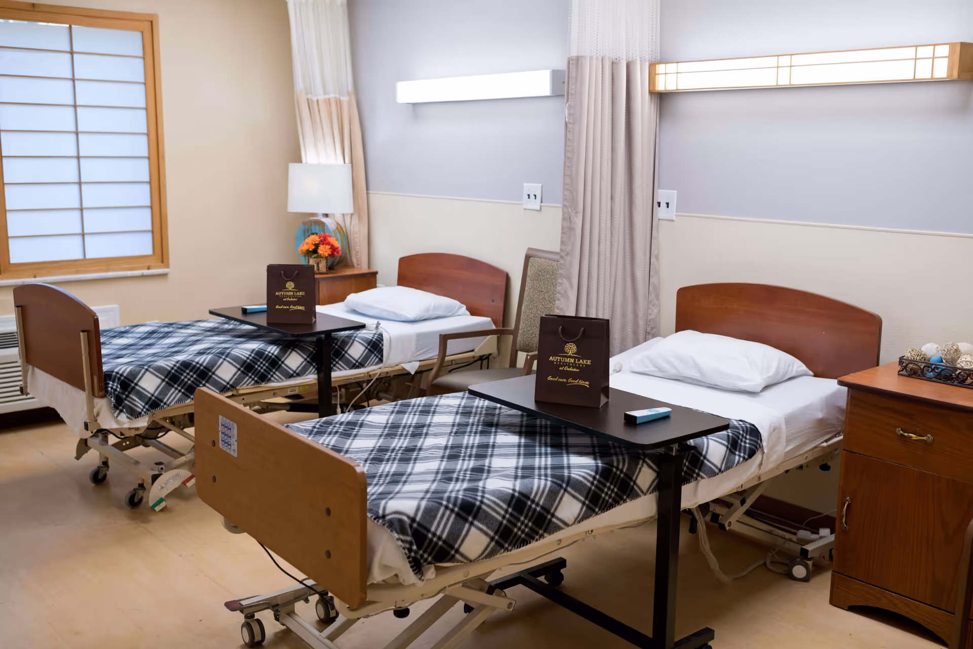 A room with two hospital-style beds, each with a plaid blanket and a pillow. Each bed has an overbed table with a small brown gift bag labeled 'Autumn Lake Healthcare at Oakview' and a remote control. There is a wooden nightstand between the beds with a lamp and a vase of flowers. The room has light-colored walls, a window with a wooden frame and shoji-style screen, and a curtain divider between the beds.