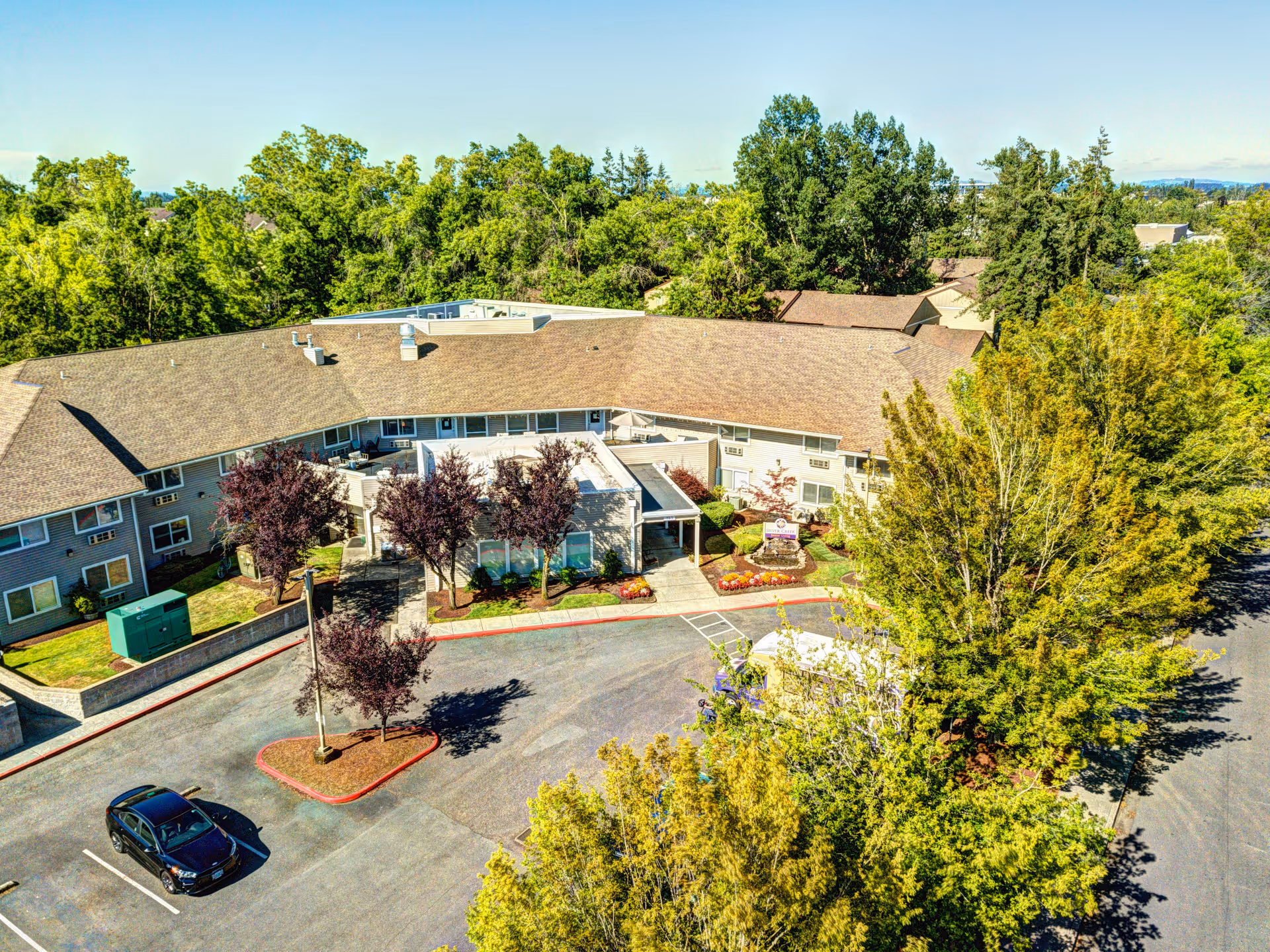 Aerial view of Silver Creek Senior Living facility showing a large two-story building surrounded by trees and greenery. There is a parking lot with a few cars and landscaped areas with small trees and shrubs near the entrance.