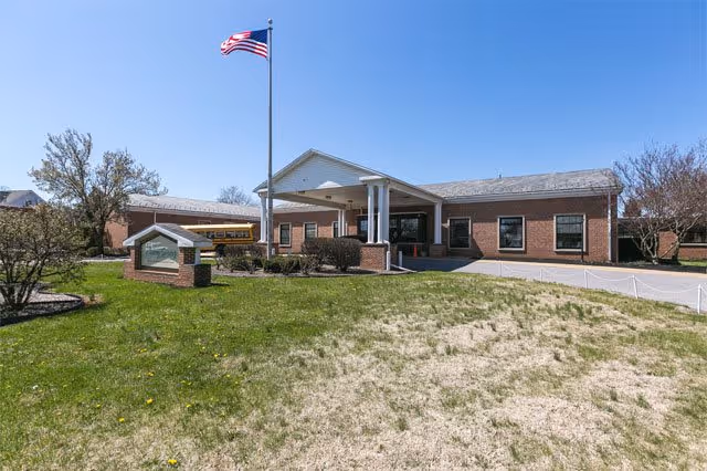 Brick single-story senior living facility front entrance with a covered portico, an American flag on a pole, and a small lawn sign.