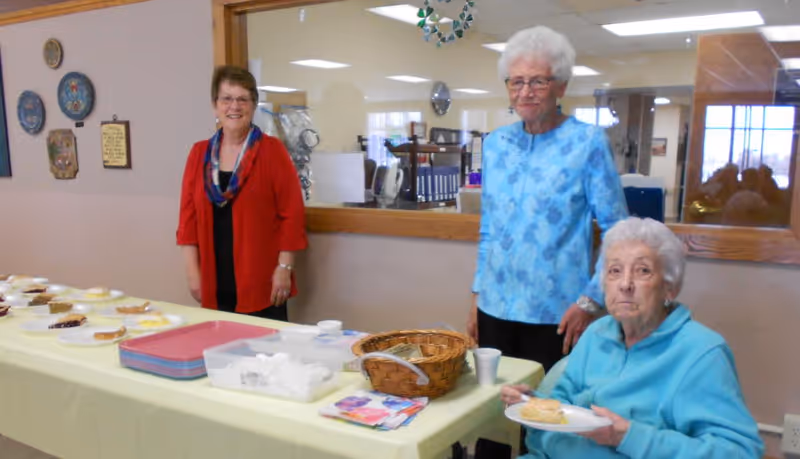 Three elderly women in a room at Norseland Nursing Home. Two women are standing behind a table covered with a yellow tablecloth, which has plates of food, trays, and a basket. One woman is seated at the table holding a plate with a sandwich. The room has a large window looking into another room with office supplies visible.