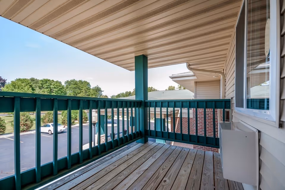 Covered wooden balcony with green railing overlooking a parking lot and trees under a clear sky.