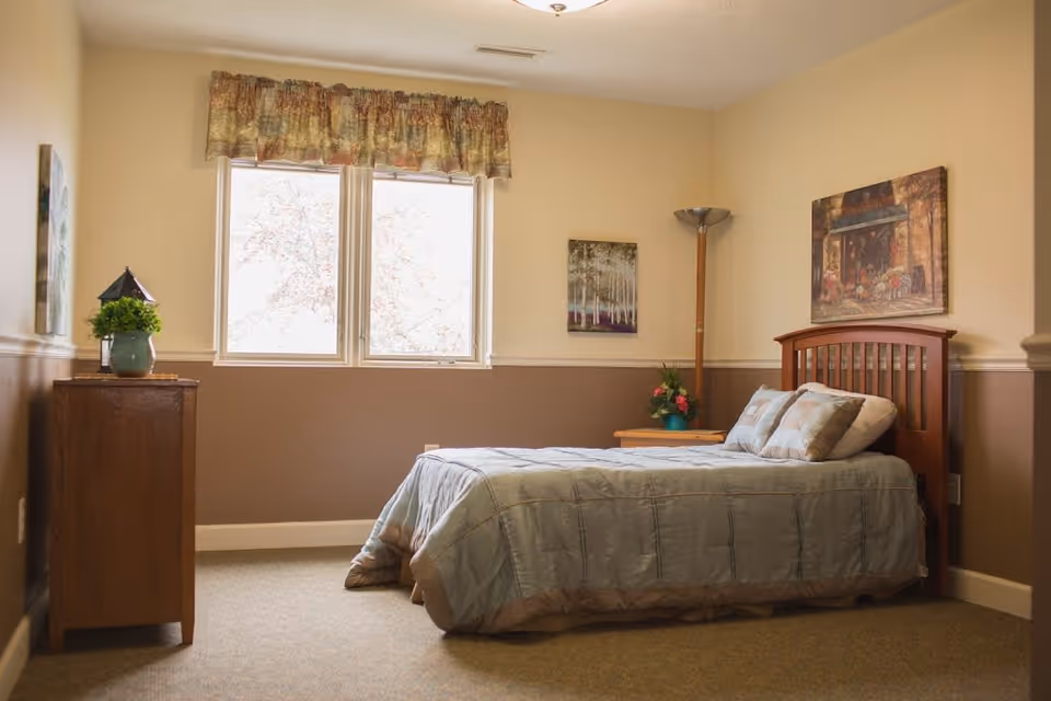 A cozy bedroom with a single bed covered in a light blue comforter and matching pillows. The room has beige and brown walls, a window with floral curtains, a wooden nightstand with a flower pot, a tall floor lamp, and framed artwork on the walls.