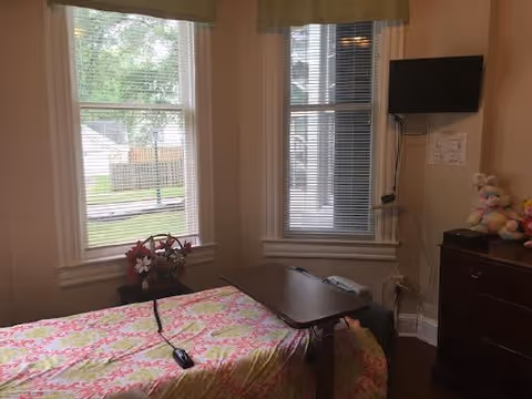A bedroom with a bed covered in a pink and green floral patterned blanket. There is a small table over the bed with a remote control on it. The room has two large windows with white blinds and beige curtains, letting in natural light. A flat-screen TV is mounted on the wall above a dark wooden dresser, which has some stuffed animals on top. Outside the windows, greenery and a fence are visible.