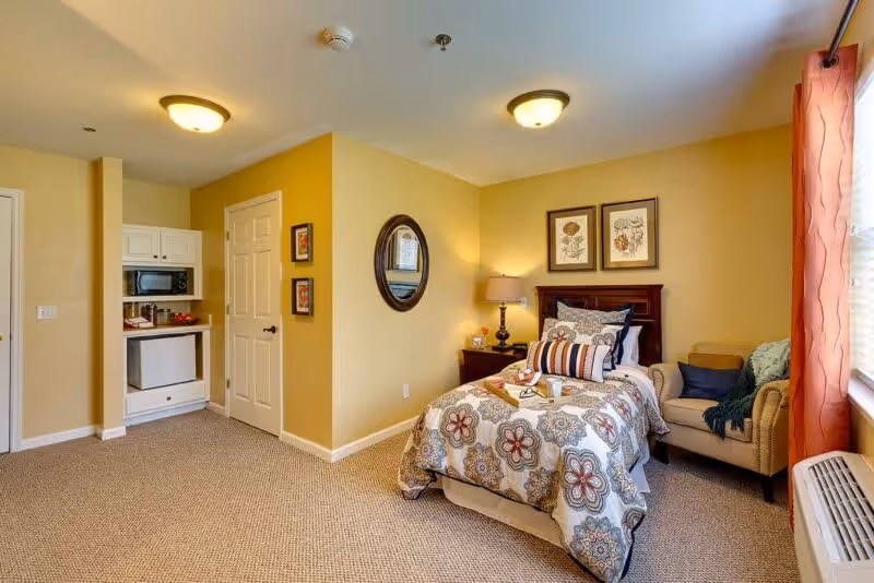A furnished bedroom featuring a single bed with patterned bedding, nightstand and armchair in a well-lit senior living suite with a small kitchenette.