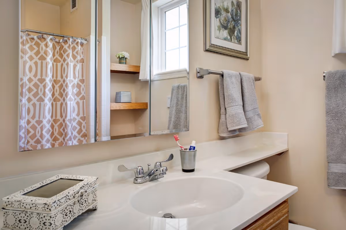 A clean bathroom with a white sink and countertop, a silver faucet, a toothbrush and toothpaste in a cup, two gray towels hanging on a towel rack, a decorative box on the counter, a window with white curtains, a framed floral picture on the wall, and a shower with a beige and white patterned curtain.