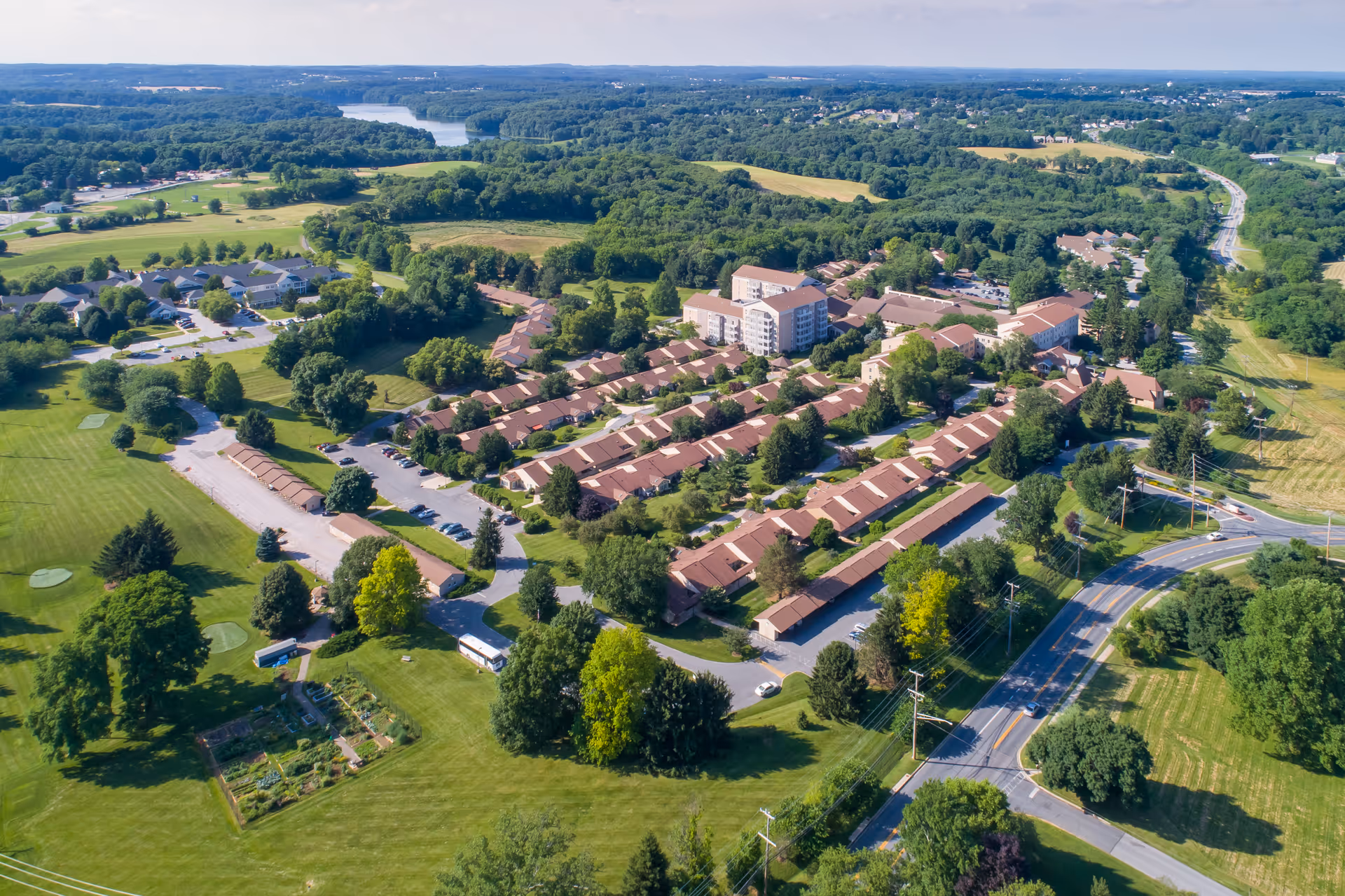 Aerial view of a large senior living facility named Fairhaven, showing multiple residential buildings with brown roofs surrounded by green lawns, trees, and roads. The facility is situated in a lush, rural area with a river visible in the distance and a golf course nearby.