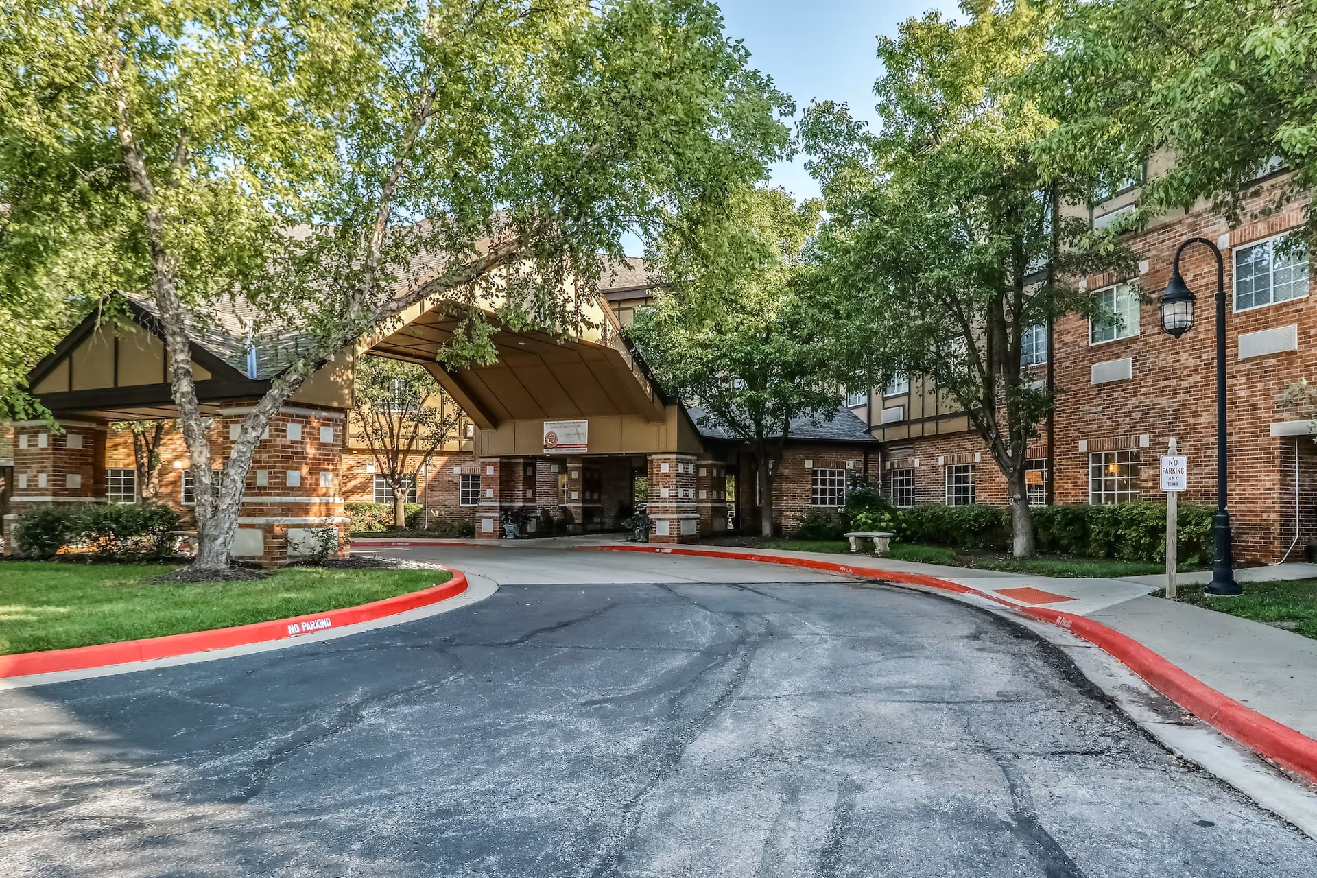 Exterior view of a senior living facility named Lamar at Town Center, showing a brick building with multiple windows, a covered entrance driveway, trees, and a sidewalk with a no parking sign.