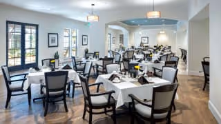 A spacious dining room with multiple tables covered in white tablecloths, each set with napkins and small flower vases. The room features wooden flooring, large windows allowing natural light, and modern light fixtures hanging from the ceiling.