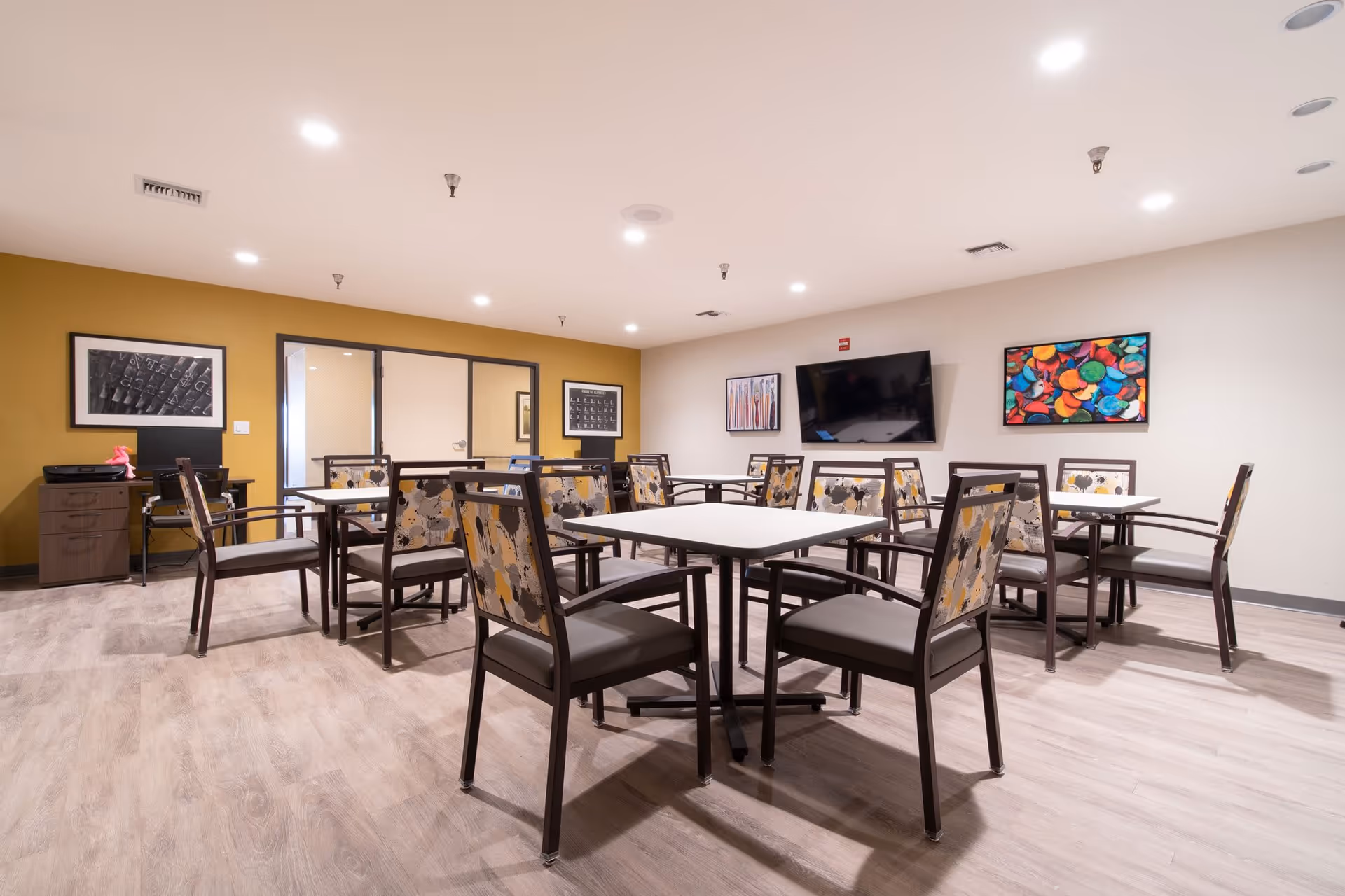 A well-lit dining room with multiple square tables and chairs featuring patterned backs. The room has light wood flooring, a mustard yellow accent wall with framed artwork, and a flat-screen TV mounted on a white wall. There is a small desk with a printer and some items on it in the corner.