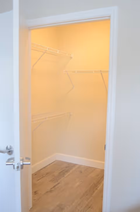 Empty walk-in closet with white wire shelving on the walls and wood-look flooring, viewed through an open white door.