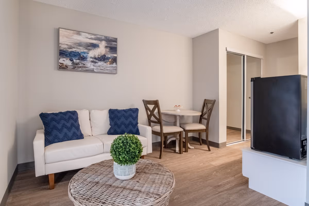 A small living area with a white couch adorned with two blue pillows, a round wicker coffee table with a green potted plant, a small round dining table with two wooden chairs, and a black mini fridge on a white stand. There is a painting of ocean waves on the wall above the couch and a sliding mirrored closet door in the background.