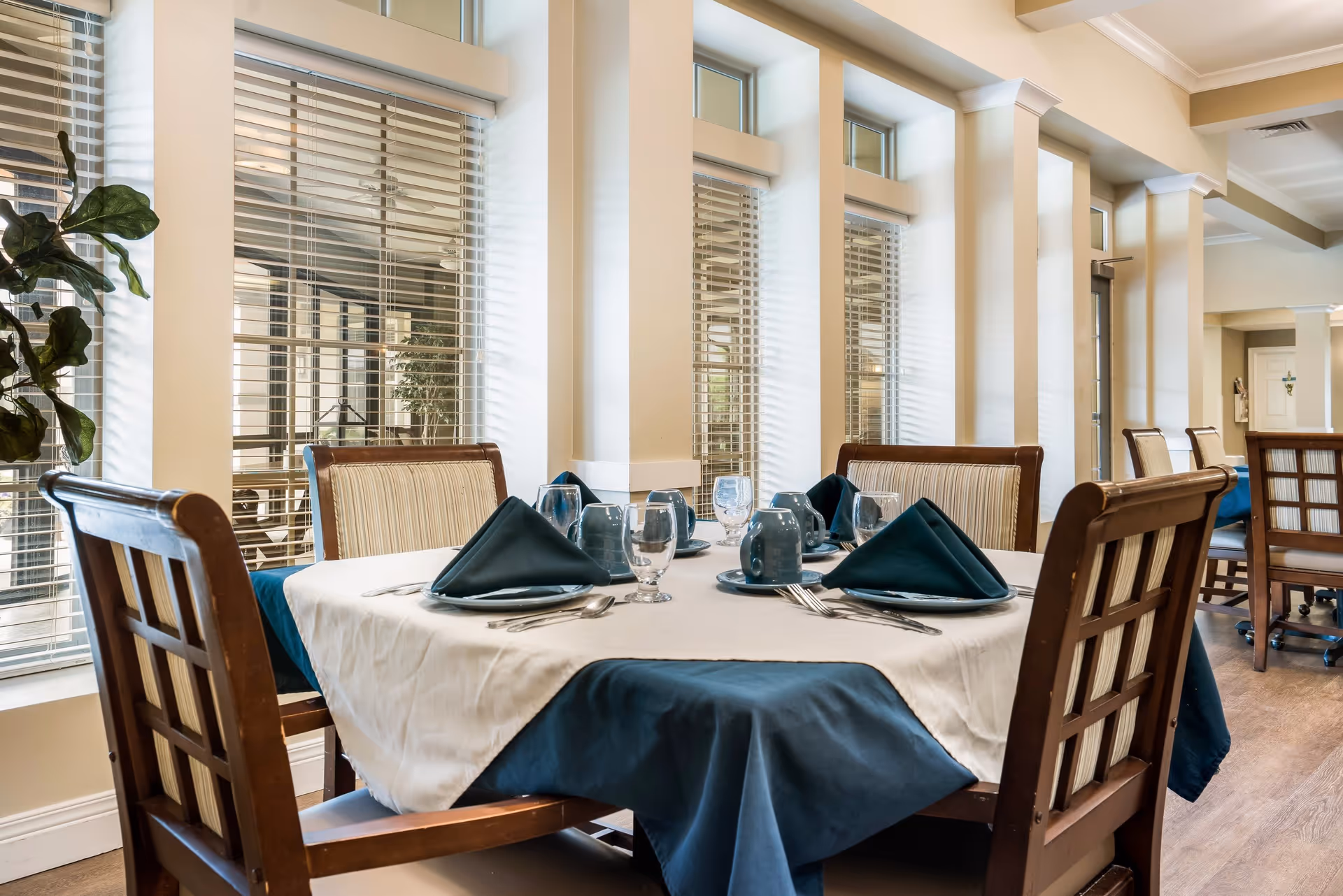 A dining table set for six with blue and white tablecloths, blue napkins folded in a triangular shape, blue mugs, water glasses, and silverware. The table is surrounded by wooden chairs with cushioned seats and backs. The room has large windows with blinds and light-colored walls, creating a bright and inviting atmosphere.