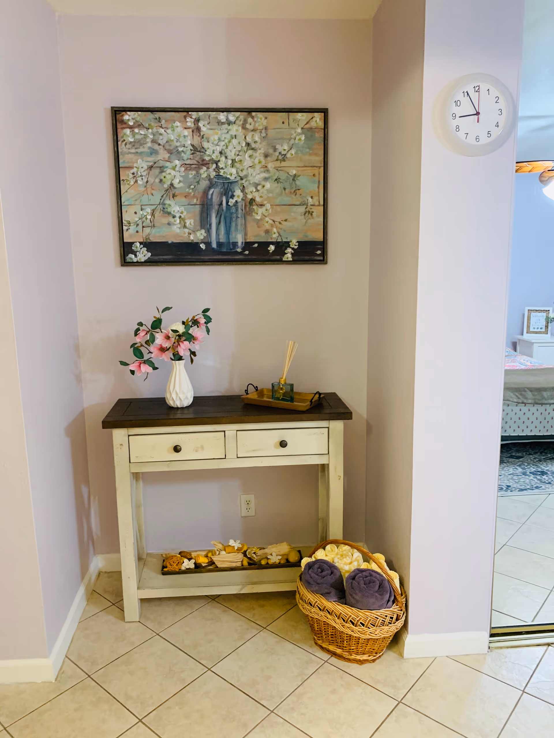 A small corner table with two drawers against a light purple wall. On top of the table is a white vase with pink flowers and a tray with a reed diffuser. Below the table are decorative items, and next to it is a wicker basket containing rolled purple towels and cream-colored items. Above the table hangs a painting of white flowers in a blue vase. A white clock showing the time as 11:50 is mounted on the adjacent wall. The floor is tiled, and a glimpse of a bedroom with a bed and framed picture is visible through an open doorway.
