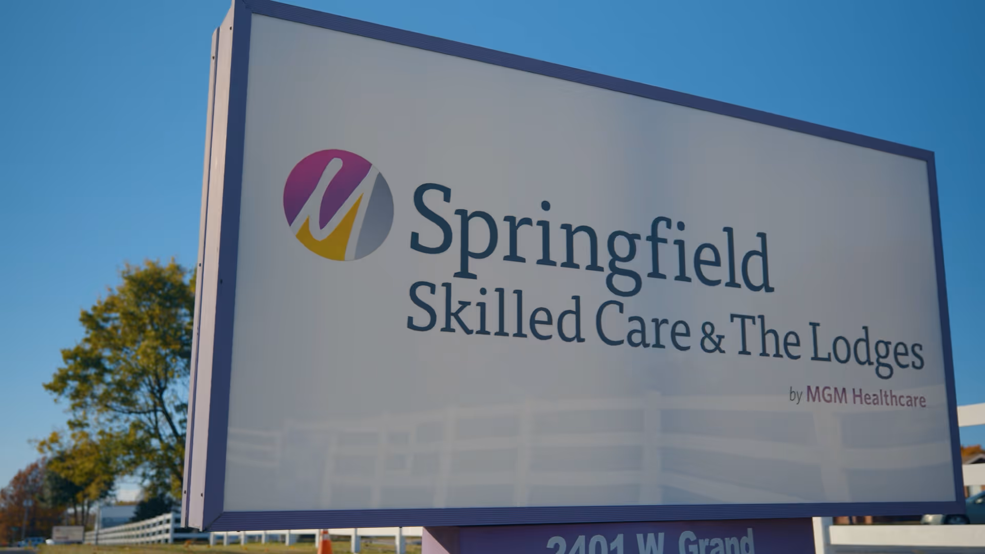Outdoor sign for Springfield Skilled Care & The Lodges by MGM Healthcare, with a clear blue sky and some trees in the background.