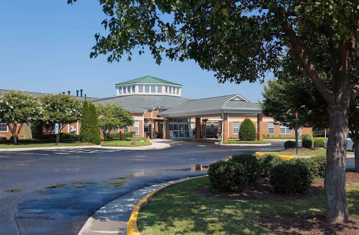 Exterior view of Dunlop House Assisted Living & Memory Care building with a circular driveway, green trees, bushes, and a clear blue sky.