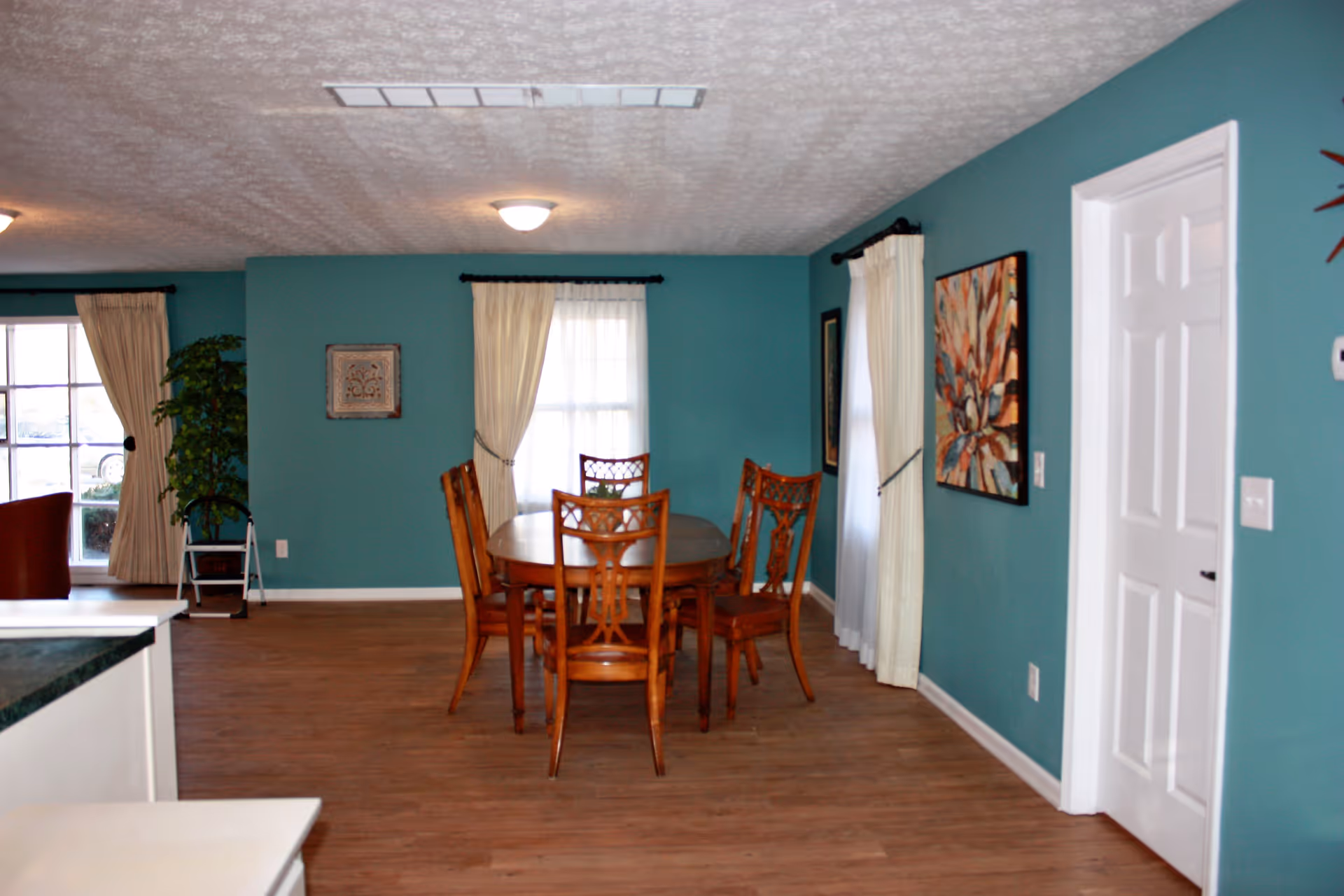 A dining area with a wooden oval table and six matching wooden chairs. The walls are painted teal and decorated with framed artwork. There are two windows with cream-colored curtains, and the floor is wooden. A white door is visible on the right side of the image.