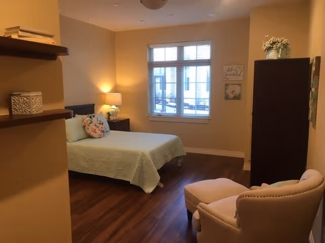 A cozy bedroom with a bed covered in a light green quilt and a floral pillow. There is a wooden nightstand with a lamp and a small plant next to the bed. A window with white blinds lets in natural light. A cushioned armchair with an ottoman is positioned near the corner. The room has wooden flooring and neutral-colored walls with some decorative wall art and shelves.