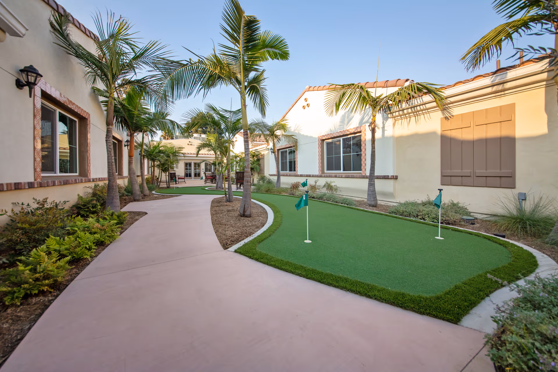 Outdoor courtyard area at Regency Palms Oxnard featuring a putting green with small flags, palm trees, and a paved walkway surrounded by beige buildings with windows and decorative shutters under a clear blue sky.