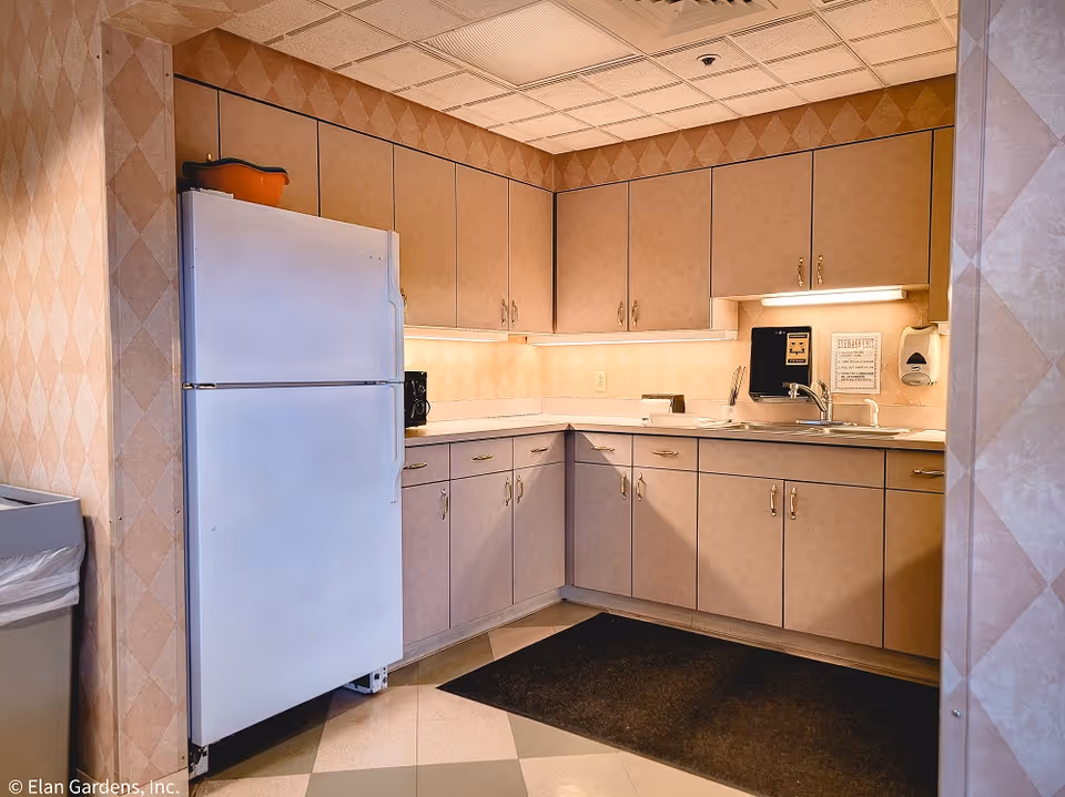 A small kitchen area with beige cabinets and a white refrigerator. The countertop has a sink, a coffee maker, and some utensils. The walls have a diamond patterned wallpaper, and there is a black mat on the floor in front of the sink. Under-cabinet lighting illuminates the countertop.