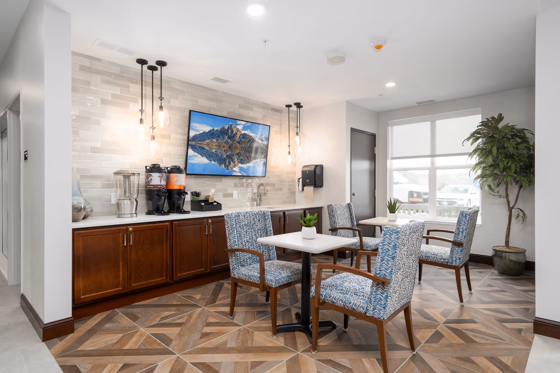 A bright and modern common area with two small square tables, each surrounded by four blue patterned upholstered chairs with wooden arms. Behind the tables is a counter with dark wood cabinets, a sink, two beverage dispensers, and a water pitcher. Above the counter is a large flat-screen TV mounted on a tiled wall, displaying a mountain landscape. Pendant lights hang from the ceiling, and a large window with white blinds lets in natural light. A tall potted plant is in the corner near the window.