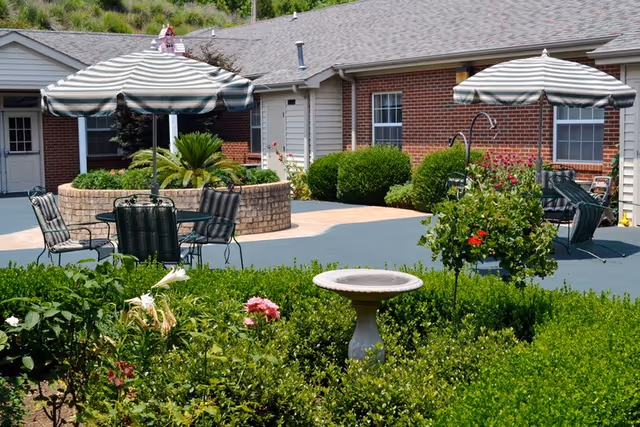 Outdoor patio area with striped umbrellas, metal chairs, and tables surrounded by green bushes and flowering plants in front of a single-story brick and siding building.
