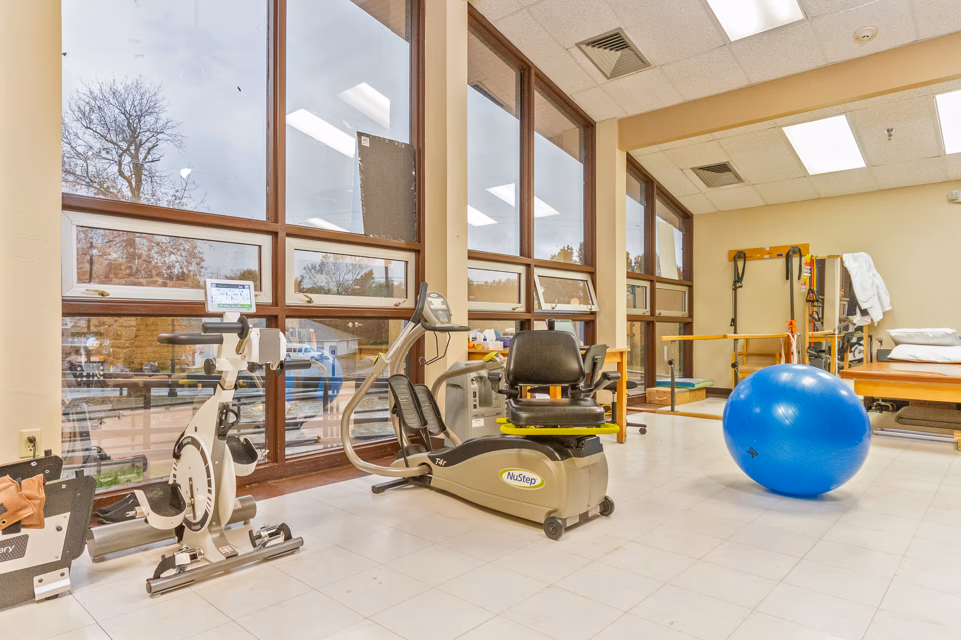 Therapy/exercise room with exercise bikes, a NuStep machine, a large blue therapy ball, and floor-to-ceiling windows.