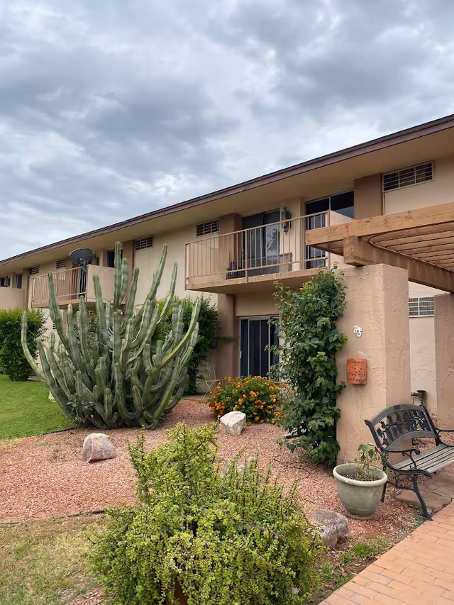 Exterior view of a two-story residential building with balconies, desert landscaping including a large cactus, bushes, and flowers, a bench, and a potted plant under a pergola structure. The sky is cloudy.