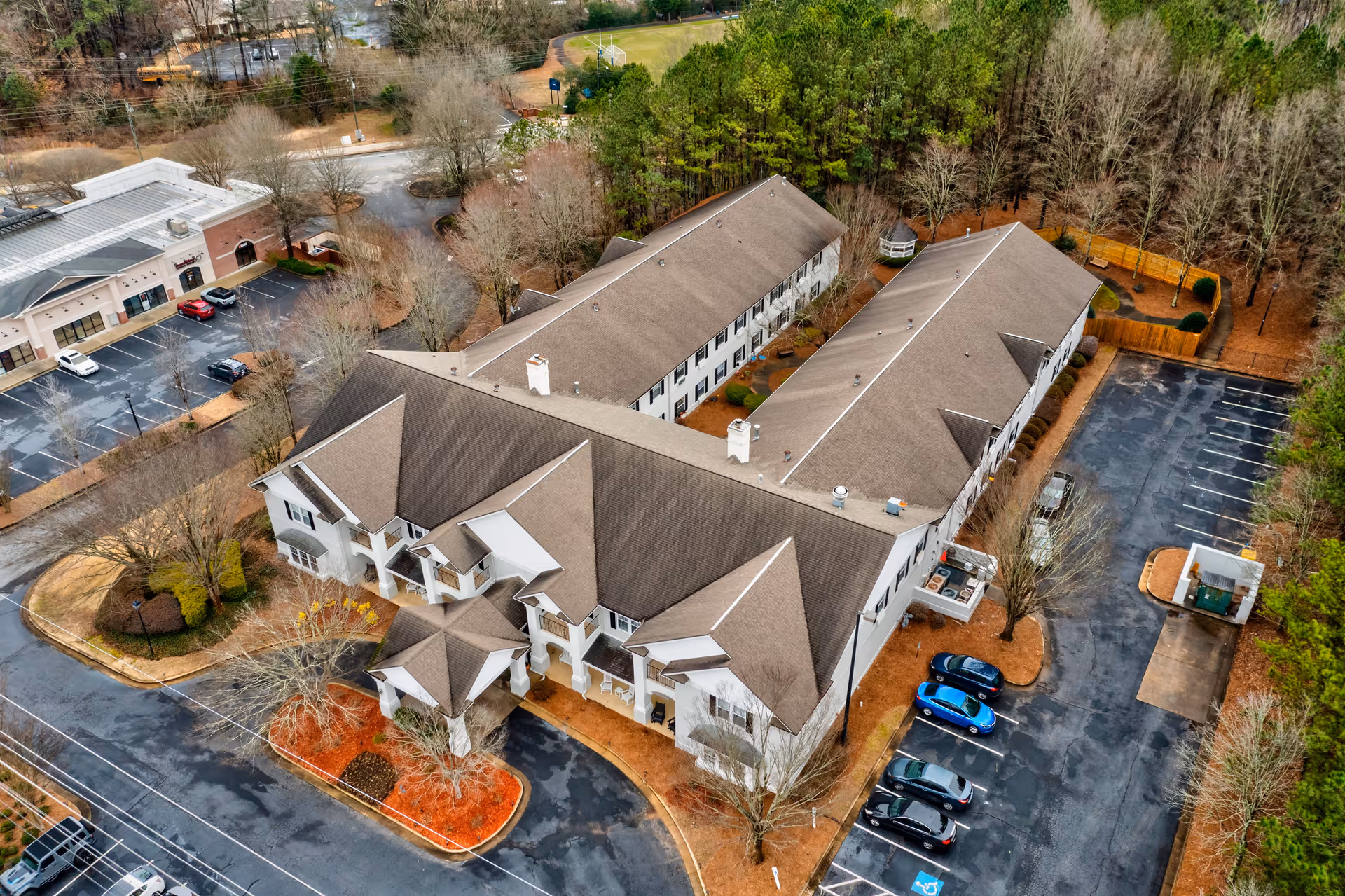 Aerial view of a large senior living facility building with a brown roof surrounded by parking lots and trees. The building has multiple wings and a covered entrance. Several cars are parked in the parking areas, and there is a small gazebo visible near the back of the property.