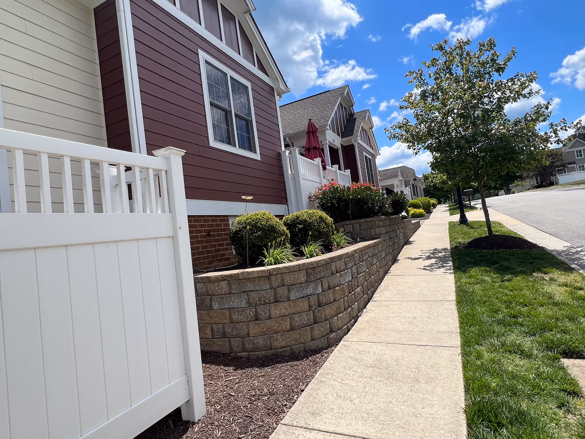 Sidewalk beside a row of red-sided townhomes with a white fence, landscaped retaining wall, and trees under a blue sky.