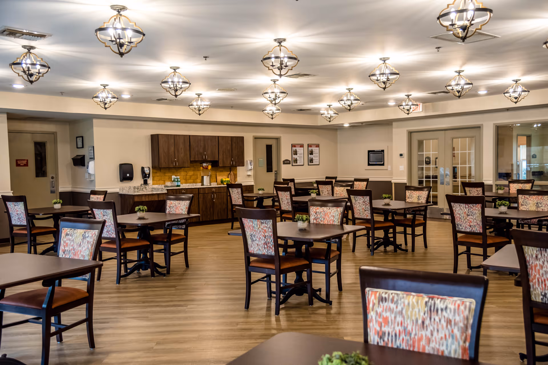 A spacious dining room with multiple dark wood tables and chairs featuring colorful patterned upholstery. The room has wooden flooring, modern ceiling light fixtures, and a counter area with cabinets and a yellow backsplash. Small potted plants are placed on each table, and there are double glass doors on one side of the room.