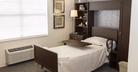 A small, neatly arranged bedroom with a single hospital-style bed covered with a white blanket. The bed has a dark brown headboard and footboard, and a small overbed table is placed on top. There is a dark brown shelving unit with a lamp, books, and framed photos next to the bed. Two framed pictures hang on the wall above the lamp. A large window with closed blinds is on the left side, and a wall-mounted air conditioning unit is below the window. The room has light-colored walls and carpeted floor.