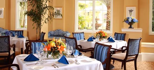 Bright dining room with round tables set with white tablecloths, blue napkins, floral centerpieces and upholstered chairs near large windows.