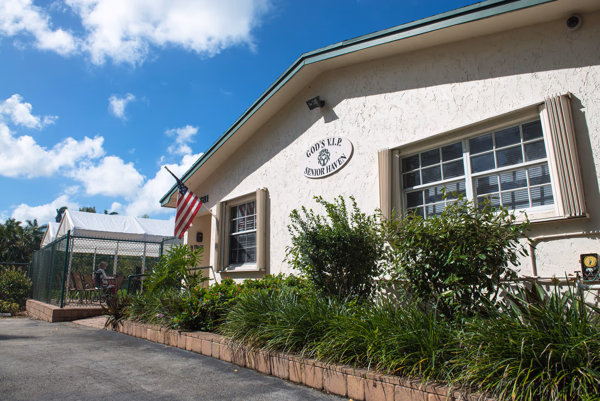 Exterior view of a single-story senior living facility building with beige stucco walls, two windows with shutters, and an American flag mounted near the entrance. There is a sign on the building that reads 'GOD'S V.I.P. SENIOR HAVEN'. The building is surrounded by green shrubs and plants, and there is a fenced outdoor seating area with chairs and a table. The sky is blue with some clouds.